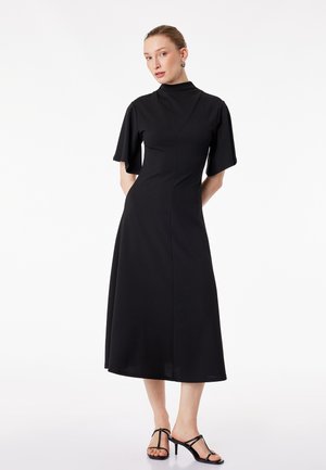 Kvinde stående iført en sort midi-kjole med flagrende ærmer, høj halsudskæring og sorte hæl-sandaler med stropper på en ensartet hvid baggrund.