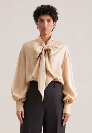 Beige satin bluse med en stor sløjfe ved halsen, ballonsleeves og en glat tekstur, parret med skræddersyede sorte bukser.