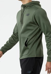 Sudadera con cremallera en tejido verde oscuro con capucha, bolsillos tipo canguro y logo negro en el pecho. Textura suave y diseño ajustado.