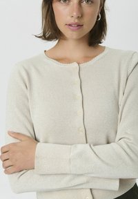Cardigan beige clair avec une texture douce, un col rond et une fermeture à boutons. Il présente un léger éclat et des poignets côtelés.