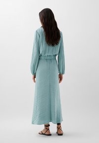 Un vestito di tessuto blu chiaro, stropicciato, con maniche lunghe e una vita elastica, abbinato a sandali neri con cinturini. Vista posteriore.