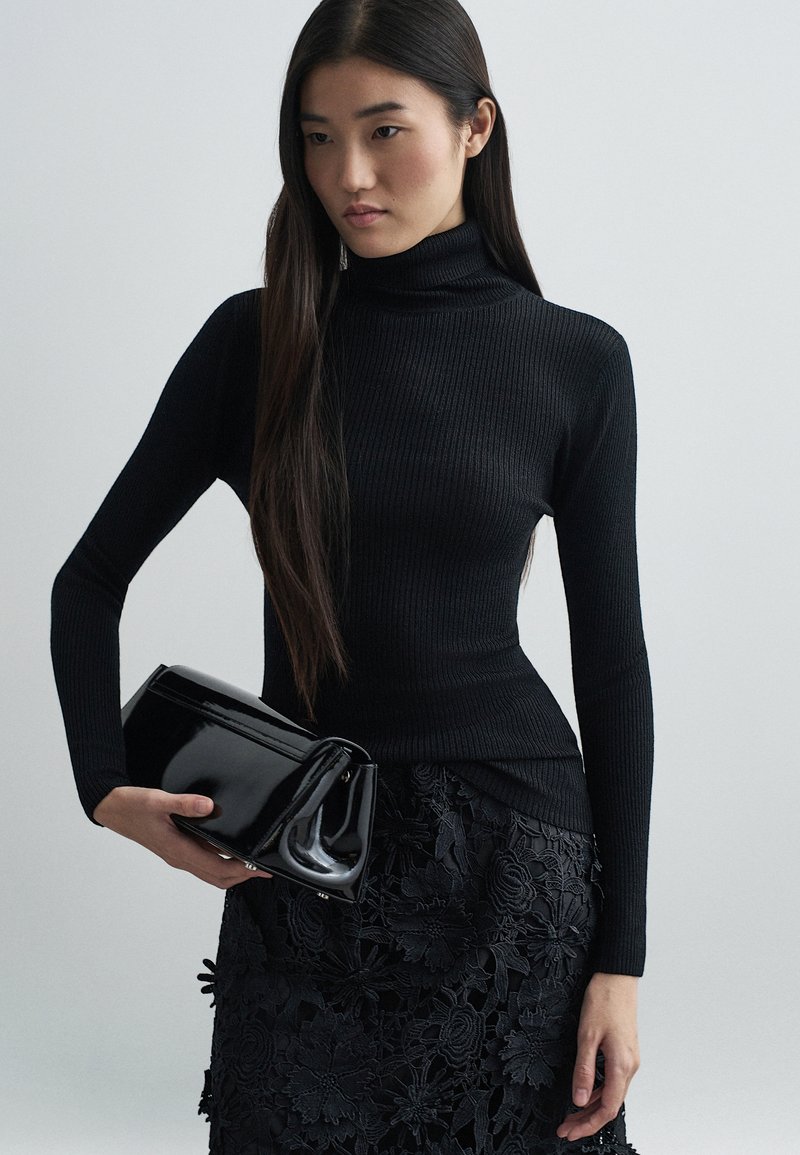 Bluză neagră cu guler înalt și textură canelată, asociată cu o fustă neagră din dantelă florală. Ține în mână o poșetă neagră lucioasă, cu un design structurat.