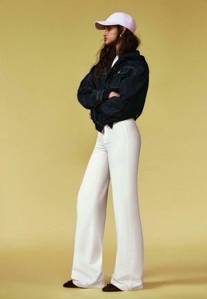 Frau steht mit verschränkten Armen, trägt eine hellrosa Mütze, eine schwarze Jeansjacke, weiße weite Hosen und schwarze Schuhe vor einem beigen Hintergrund.