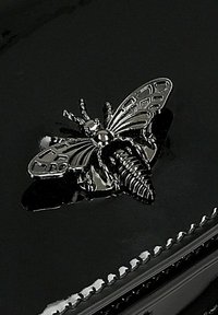 Černý lakovaný povrch s detailním stříbrným emblémem včely, který má propracovaná křídla a texturu těla, umístěný uprostřed produktu.