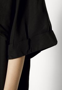 Gros plan sur le bras d’une personne portant un vêtement noir avec une manche courte retroussée sur un fond uni.