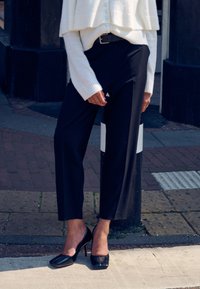 Črne hlače po meri z široko nogo in visokim pasom, kombinirane s kremnim pletenim krajšim puloverjem. Obutev vključuje sijoče črne čevlje z visokimi petami.