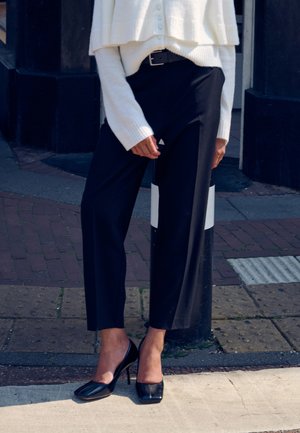 Pantaloni neri su misura, a gamba larga e vita alta, abbinati a un maglione corto lavorato a maglia color crema. Calzature con scarpe con tacco alto nere lucide.