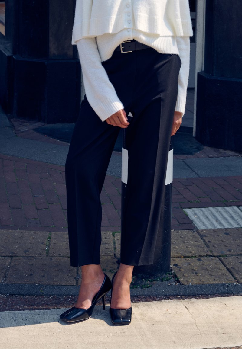 Črne hlače po meri z široko nogo in visokim pasom, kombinirane s kremnim pletenim krajšim puloverjem. Obutev vključuje sijoče črne čevlje z visokimi petami.