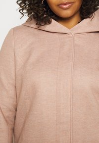 Ljusrosa huva jacka i strukturerad tyg, med en ren design och subtila sömdetaljer på framsidan.