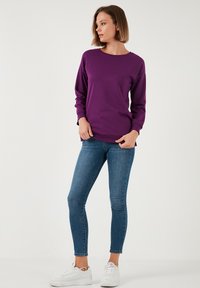 Felpa lunga viola con collo a giro, abbinata a jeans skinny blu e sneakers bianche su uno sfondo chiaro.