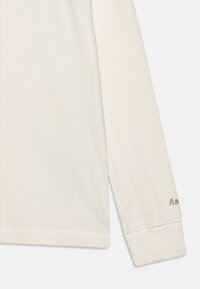 Long-sleeve cream top made of ribbed fabric, featuring a rounded neckline and small embroidered branding in brown on the sleeve.