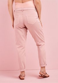 Pantalones de mezclilla rosa claro con un diseño ajustado, dobladillos enrollados y dos bolsillos traseros. Llevados con sandalias planas, sobre un fondo rosa pastel.