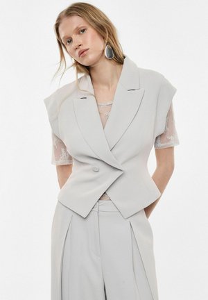 Donna che indossa blazer senza maniche su misura grigio chiaro sopra top in pizzo floreale trasparente e pantaloni a vita alta a gamba larga coordinati, su sfondo bianco.