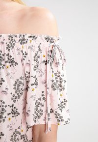 Top con spalle scoperte in tessuto leggero rosa con motivi floreali, caratterizzato da un colletto arricciato e dettagli con fiocco sulle maniche.