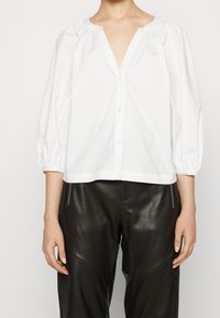 Weiße Bluse mit langen Ärmeln, V-Ausschnitt und gerafften Details an den Schultern. Hergestellt aus leichtem Baumwollstoff, mit Knopfverschluss.