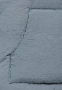 Hellblaues Gewebe mit texturierter Oberfläche, ausgestattet mit einer aufgesetzten Tasche mit sichtbaren Nähten. Minimales Design und weicher, zerknitterter Look.