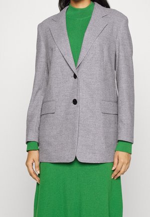 Personne portant un blazer gris clair à deux boutons sur une robe en maille côtelée verte à manches longues, debout devant un fond uni.