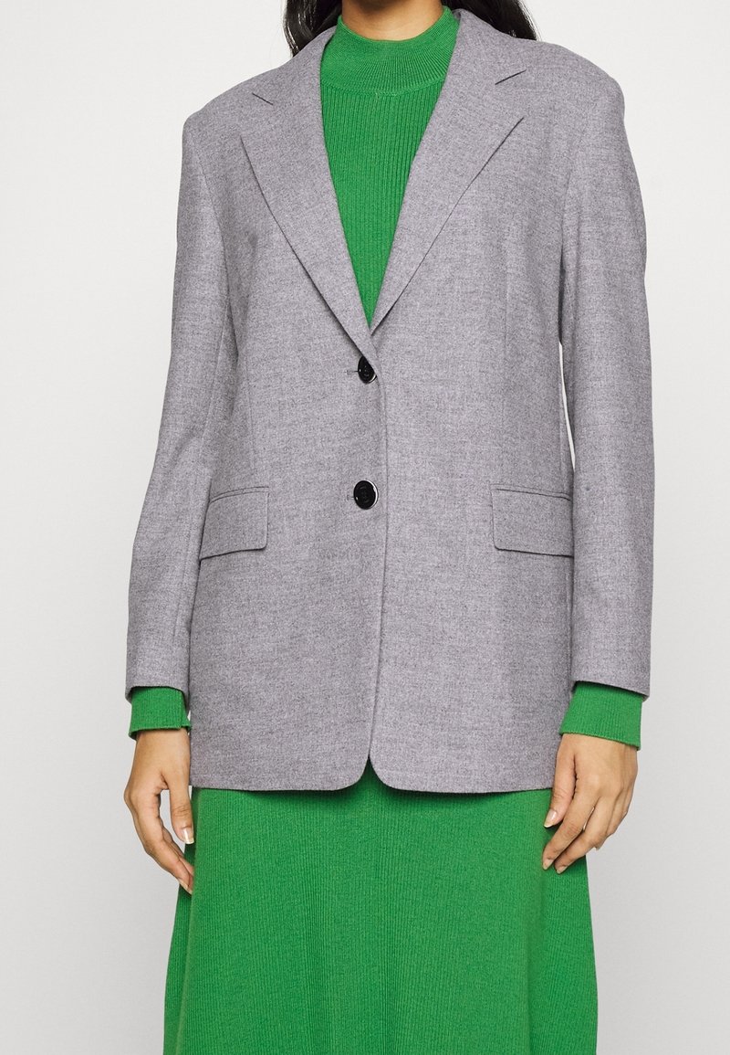 Personne portant un blazer gris clair à deux boutons sur une robe en maille côtelée verte à manches longues, debout devant un fond uni.