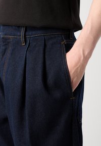 Mørkeblå denimbukser med plisseringer, der har sidelommer og kontrastfarvet orange syninger. Glat tekstur med en skræddersyet pasform.