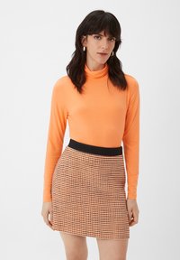 Oranje langlang mouw turtleneck top gecombineerd met een zwarte tailleband mini-rok met een houndstoothpatroon in oranje en zwart.