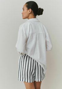 Camicia bianca, ampia, con scollo a colletto e maniche arrotolate, abbinata a pantaloncini a righe grigie e bianche in tessuto leggero.