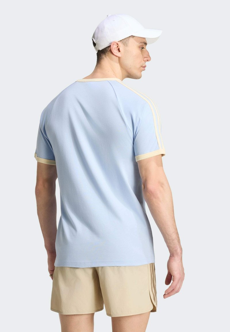 Uomo che indossa una maglietta azzurro chiaro con rifiniture beige, pantaloncini beige e cappellino bianco, in piedi e guarda di lato su uno sfondo semplice.