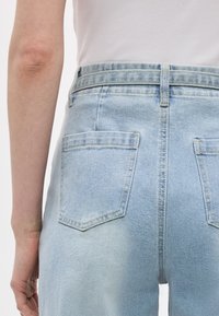 Άτομο που φοράει ανοιχτό μπλε τζιν με ψηλή μέση και λευκό ριμπ αμάνικο τοπ, εμφανιζόμενο από πίσω και από τη μέση και πάνω.