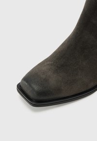 Stivale alla caviglia in suede grigio con punta quadrata e suola nera piatta, caratterizzato da una superficie liscia e dettagli minimi.