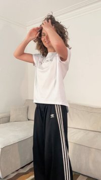 Bela majica s tekstom, crne trenirke s tri bijele pruge; kovrčava kosa; unutarnji ambijent s kaučem u pozadini.