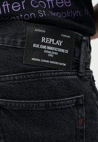 Sorte denimjeans med en rektangulær sort lap, der viser teksten "REPLAY Blue Jeans Manufacturing Co Established 1981", båret sammen med en sort skjorte.