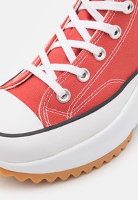 Baskets en toile rouge avec œillets et lacets blancs, bout en caoutchouc, bordure noire contrastante et semelle en gomme texturée avec un motif en zigzag.