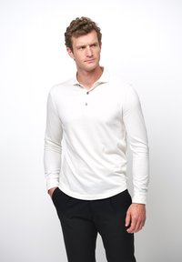 Langarm-Polohemd aus weißer Baumwolle, mit einer Knopfleiste mit drei Knöpfen und einem abgerundeten Saum. Kombiniert mit schwarzen Hosen.