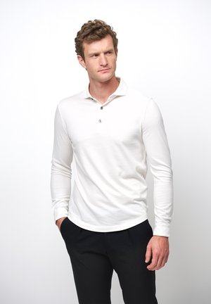 Camisa polo de manga larga en algodón blanco, con un cuello de tres botones y un dobladillo curvo. Combinada con pantalones negros.