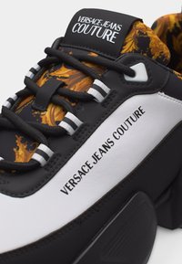 Ασπρόμαυρο αθλητικό παπούτσι Versace Jeans Couture με μαύρα κορδόνια, γλώσσα με χρυσό λουλουδάτο μοτίβο και λογότυπα στη γλώσσα και την πλευρική επιφάνεια.