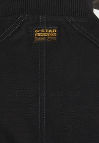 Tissu en denim noir avec une étiquette de marque dorée et noire "G-STAR ORIGINALS RAW DENIM" cousue au-dessus de la couture centrale arrière.