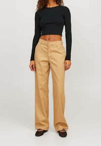 Kvinna i beige vida byxor, svart kortärmad topp och svarta mesh-balletskor mot en ljus enfärgad bakgrund.