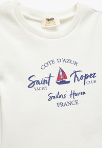 Camiseta de algodón blanca con un gráfico de un velero en azul marino y rojo, acompañado del texto: "COTE D'AZUR Saint Tropez Yacht Club Sailor's Haven Francia."