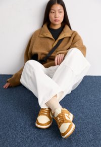 Sneakers med en kombination av tan läder och gräddfärgad mocka, med perforerad tå, vit mellansula och bruna skosnören, som bärs med vita byxor.