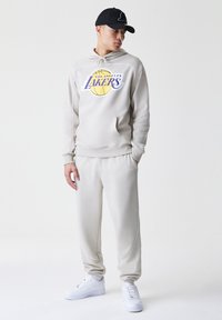 Κρέμ φούτερ με λογότυπο "Los Angeles Lakers", τσέπη καγκουρό, ταιριαστές κρέμ φόρμες και λευκά αθλητικά παπούτσια, φορεμένο με μαύρο καπέλο.