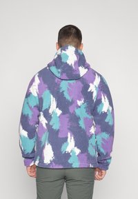 Fialová, tyrkysová a biela vzorovaná fleecová mikina s vysokým golierom a priestrannou kapucňou. Mäkká textura, voľný strih.