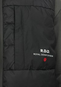 Crni prsluk s umetkom, s patentnim zatvaračem, bijelim logotipom i crvenim detaljima. Glatka vanjska tekstura s lepršavim dijelovima sprijeda.