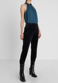 Top senza maniche turchese con scollo arricciato e bordatura decorata, abbinato a pantaloni a sigaretta in velluto scuro e stivali con tacco neri lucidi.