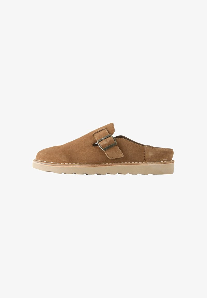 Chaussure en daim marron à enfiler avec une sangle à boucle latérale et une semelle en caoutchouc beige, conçue pour un usage décontracté et un confort facile à enfiler.
