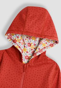 Felpa rossa con texture a pois; presenta una fodera con motivo floreale nella cappuccio. Chiusura a zip lungo la cucitura frontale.