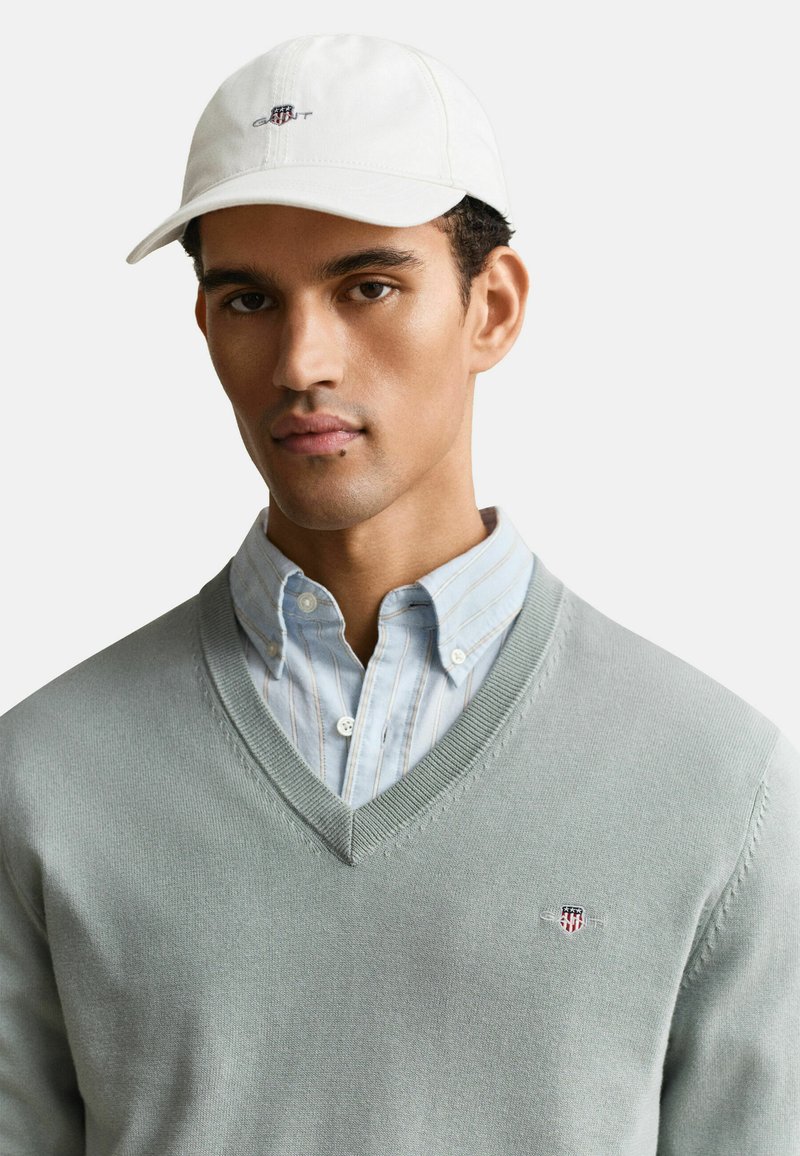 Hombre que lleva una camisa de rayas azul pálido abotonada debajo de un suéter de cuello en V gris claro y una gorra blanca de béisbol con logos bordados.