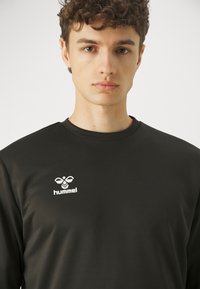 Ung mand med krøllet brunt hår iført en sort hummel langærmet trøje med hvidt logo på en ensfarvet lys baggrund.