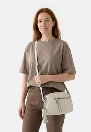 Woman with red hair stands wearing a brown t-shirt and pants, holding a beige crossbody bag with a front zipper and tassel detail.