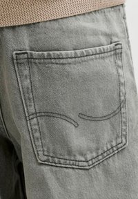 Hallkaader halli teksapükste tagatasku lähedalt, millel on topeltõmbluse detail, ja kohal on osa beežist kootud kampsunist.