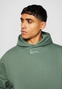 Sudadera verde caqui con un tejido texturizado, corte holgado y un logo blanco bordado en el pecho. Cuenta con una gran capucha blanda con cordón.