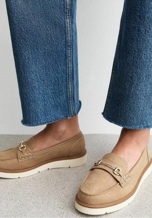 Voeten die beige suède loafers dragen met witte zolen en metalen detail, gecombineerd met blauwe cropped spijkerbroeken tegen een effen achtergrond.